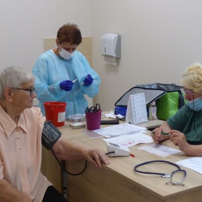 Trzy osoby. Seniorka ma mierzone ciśnienie, pielęgniarka przygotowuje szczepionkę.