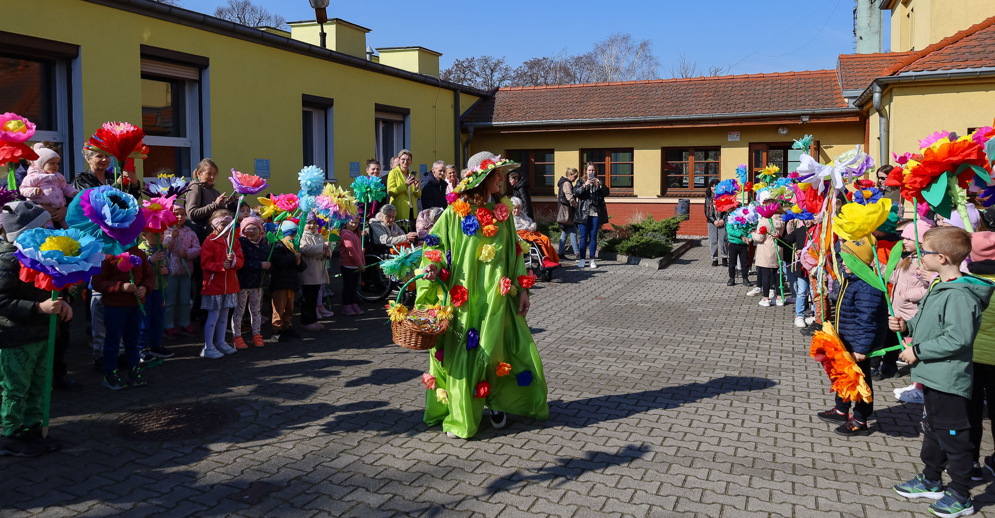 Na zdjęciu widać wydarzenie na świeżym powietrzu, które odbywa się przed budynkiem z żółtymi ścianami. Na pierwszym planie widoczna jest postać ubrana w zielony strój ozdobiony kolorowymi kwiatami. Ma na sobie kapelusz, a w ręce trzyma koszyk. Jest to prawdopodobnie osoba przebrana za wiosnę, biorąca udział w symbolicznym przywoływaniu tej pory roku. W tle widać grupę dzieci trzymających w rękach duże, kolorowe kwiaty wykonane z papieru. Wydają się być uczestnikami wiosennego wydarzenia, prawdopodobnie związanego z obchodami pierwszego dnia wiosny. Na twarzach uczestników i widzów widać uśmiechy, co świadczy o radosnym charakterze tego spotkania.